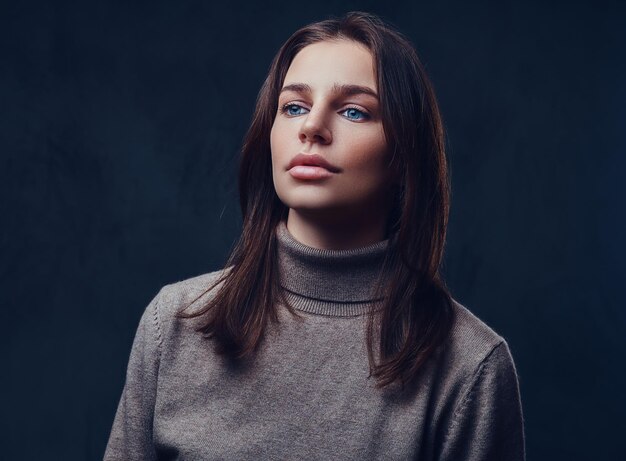 An attractive brunette female dressed in a long neck brown jacket over grey background.