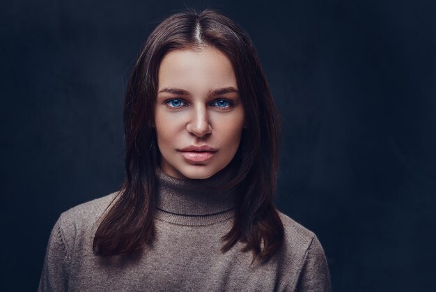 An attractive brunette female dressed in a long neck brown jacket over grey background.