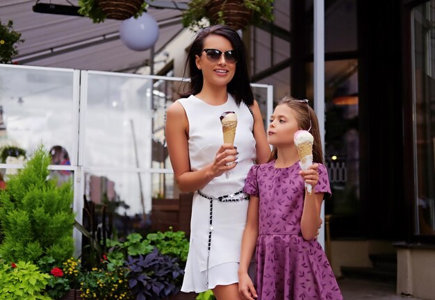 An attractive brunette female dressed in a white dress and sunglasses and cute teenage girl eating ice cream on a street.