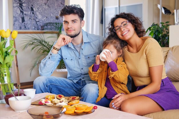 Attractive brunette female in eyeglasses, bearded stylish male and their cute little daughter sits on a couch and eat lunch in a living room.