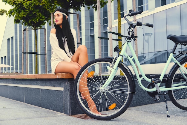 Attractive brunette female with barefoot sits on a bench over modern building background after bicycle ride.