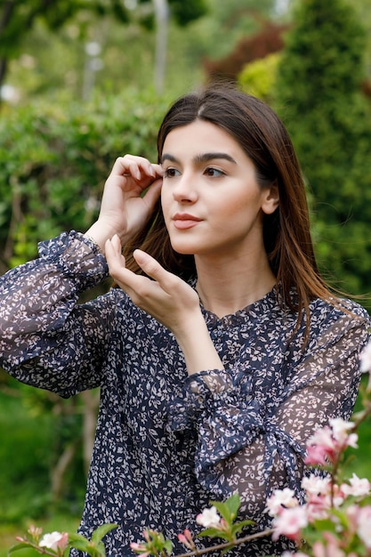 Attractive brunette girl standing in blooming garden looking forward touching hair