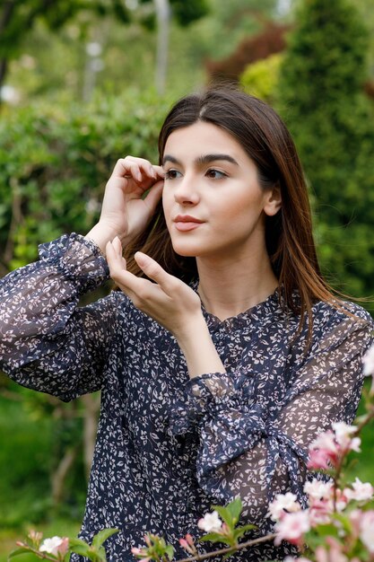 Attractive brunette girl standing in blooming garden looking forward touching hair