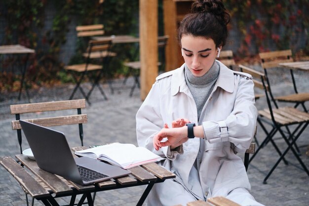 Attractive brunette girl in trench coat confidently using smartwatch studying in cafe on city street