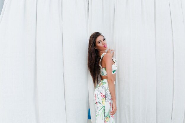 Attractive brunette girl with long hair is posing near long white curtains outdoor. She is looking to the camera.