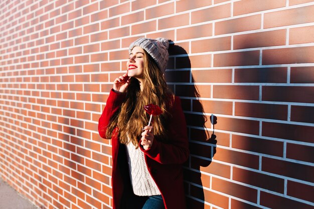 Attractive brunette girl with long hair on wall  outside. She wears knitted hat, red coat. Holds lollipop red lips. Smiling with closed eyes to sunshine.