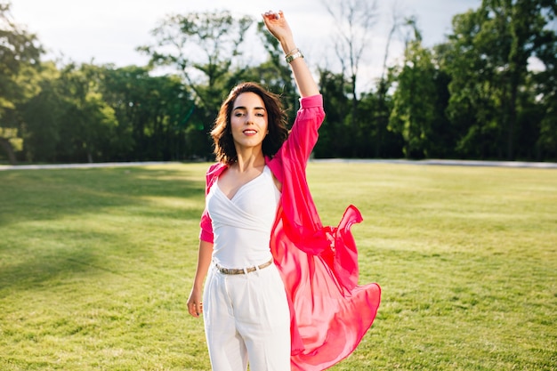 Attractive  brunette girl with short hair is walking in summer park. She wears white clothes, her long pink shirt is flying on wind. She keeps hand above and looks to the camera.