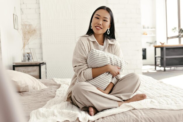 베이지색 카디건과 바지를 입은 매력적인 브루네트 여성은 카메라를 바라보고 침실에서 베개를 껴안고 세련된 옷을 입은 젊은 아시아 여성이 부드러운 침대에 앉아 있습니다.