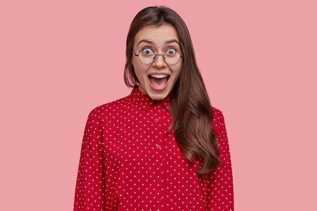 Free photo attractive brunette overjoyed woman opens mouth and yells from joy, wears pink polka dot blouse, spectacles, glad with her success