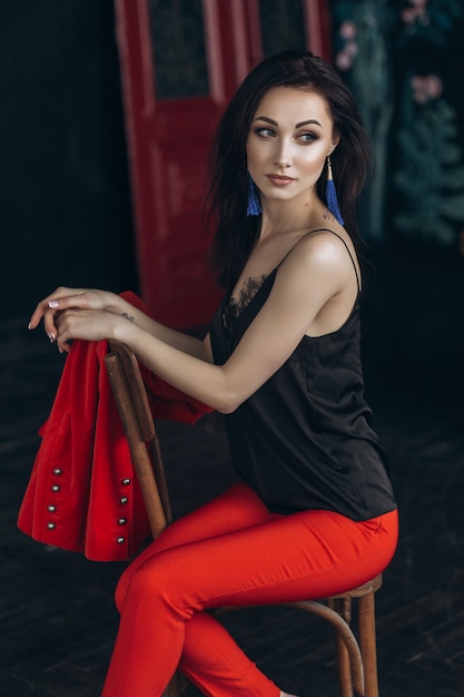 Attractive brunette in red suit sits on the old chair in dark studio