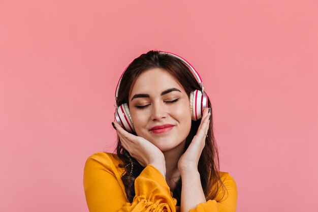 Attractive brunette with smile and eyes closed listening to song in headphones. Lady in bright yellow blouse posing on isolated wall.