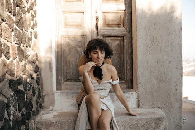 Attractive brunette woman in beige outfit sits near old house Young lady in summer dress with straw hat and bag poses near wooden door