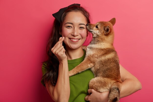 Attractive brunette woman with eastern appearance, holds Shiba Inu dog on hands, makes korean like sign, expresses love to pet, adopts animal