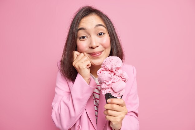 Attractive brunette young Asian woman with delighted expression holds tasty cone ice cream in waffle keeps hand on face has sweet tooth isolated over pink  wall. Yummy summer dessert