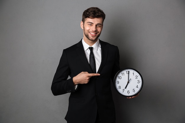 Attractive businessman in classic black suit pointing with finger on big clock, 