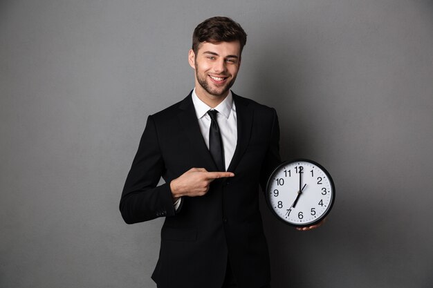 Привлекательный бизнесмен в классическом черном костюме, указывая пальцем на большие часы,