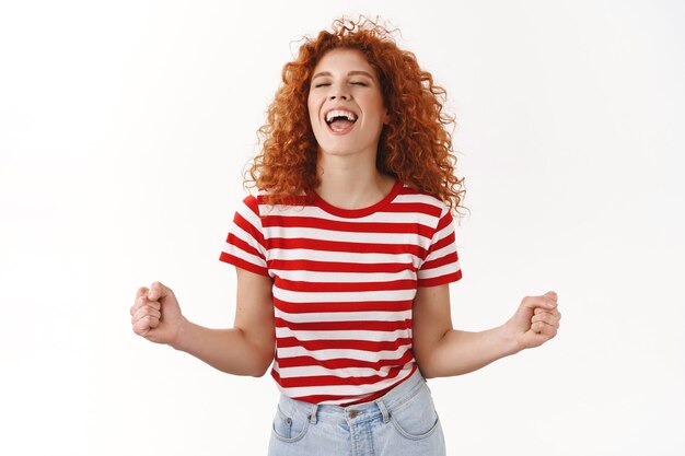 Attractive carefree lively redhead curly woman enjoy having fun release emotions dancing close eyes scream yes thrilled clench fists triumphing winning lottery