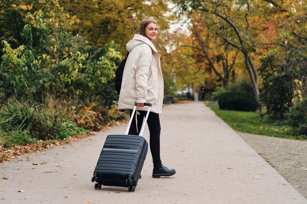 屋外のカメラで幸せそうに見ているスーツケースとダウンジャケットの魅力的なカジュアルな女の子