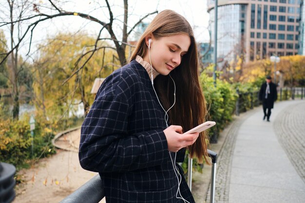 Attractive casual girl in earphones happily listening music on cellphone during walk in city park