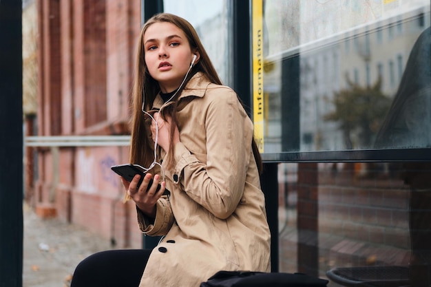 Привлекательная случайная девушка в наушниках с мобильным телефоном задумчиво ждет общественный транспорт на автобусной остановке под открытым небом