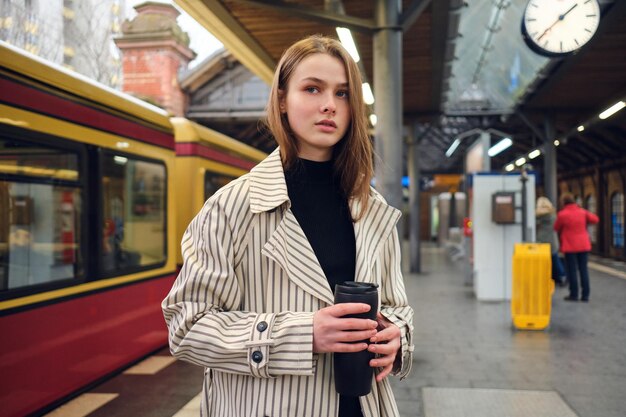 Attractive casual girl in trench coat with thermo cup waiting train at subway station