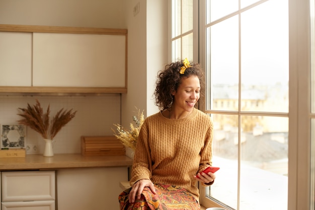 Free photo attractive casully dressed young latin woman sitting on windowsill in kitchen, holding cell phone checking newsfeed via social network or typing text message, having rest. technology and communication