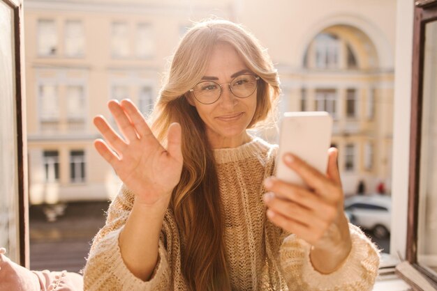 Attractive caucasian adult lady is sitting on window sill in room holding mobile waving her hand at phone camera Technology and communications