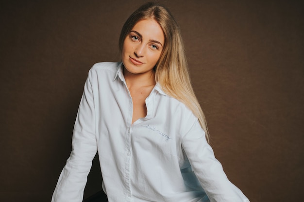 Бесплатное фото Привлекательная кавказская блондинка в белой рубашке позирует на коричневой стене