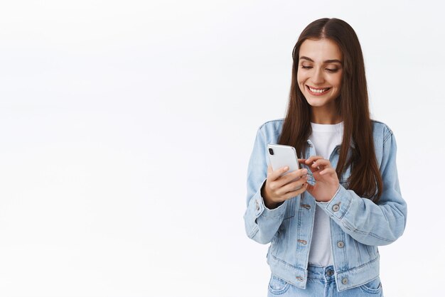 Привлекательная кавказская брюнетка, использующая приложение для смартфона и улыбающаяся, печатая что-то на мобильном дисплее, наслаждаясь просмотром социальных сетей, стоя на белом фоне, подключается к друзьям Wi-Fi