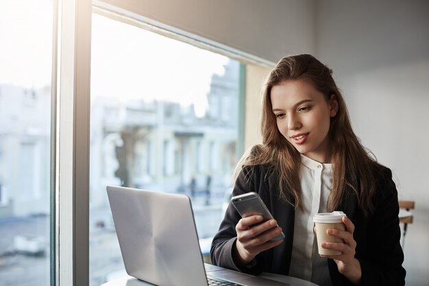 attractive caucasian businesswoman sitting in cafe near window, drinking coffee and messaging via smartphone, working with laptop during break