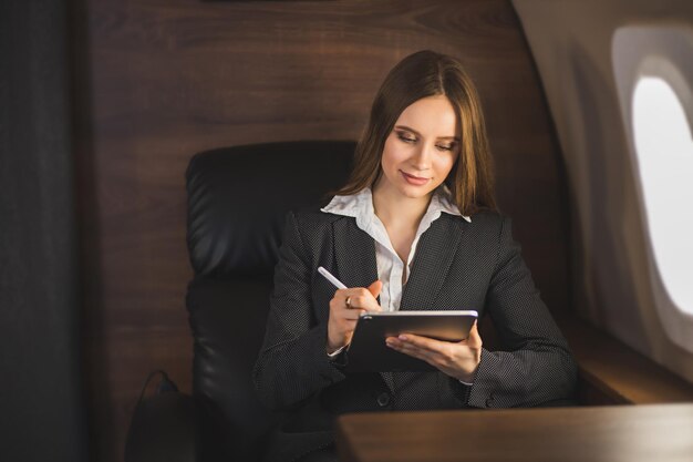 飛行機の快適な座席に座って、現代のタブレットを持って、機内でワイヤレス接続を使用して作業している魅力的な白人実業家。ビジネス、旅行、テクノロジー。