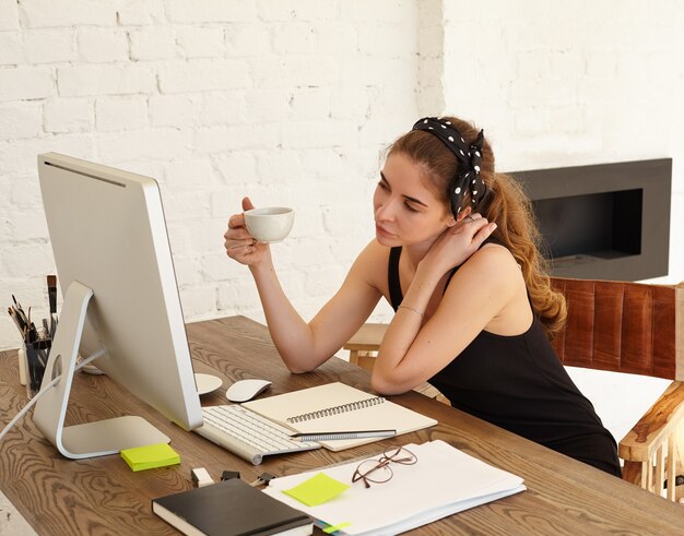 Attractive Caucasian female student playfully looks in the monitor, talking to the friend by video conference, drinking coffee. Pretty girl wearing headscarf is interested looks in the screen PC