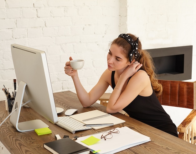 Attractive Caucasian female student playfully looks in the monitor, talking to the friend by video conference, drinking coffee. Pretty girl wearing headscarf is interested looks in the screen PC