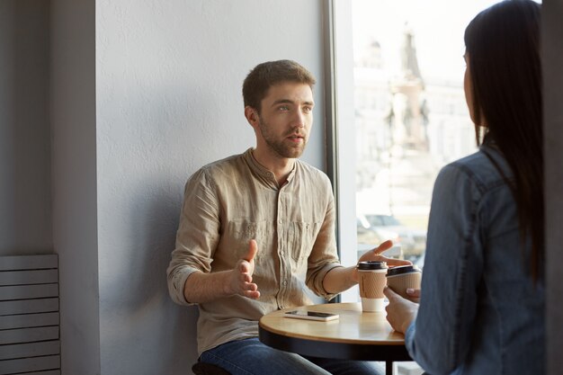 Attractive caucasian guy with dark hair and bristle sitting in cafe on a date talking to his girlfriend about his work, gesticulating with hands and drinking coffee.