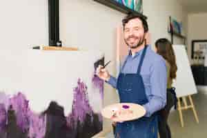 Foto gratuita uomo caucasico attraente che sorride e tiene in mano un pennello mentre è in piedi davanti alla tela che sta iniziando a dipingere per una lezione d'arte