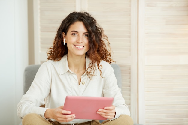 Attractive caucasian woman with curly hair wearing white shirt holding pink tablet computer