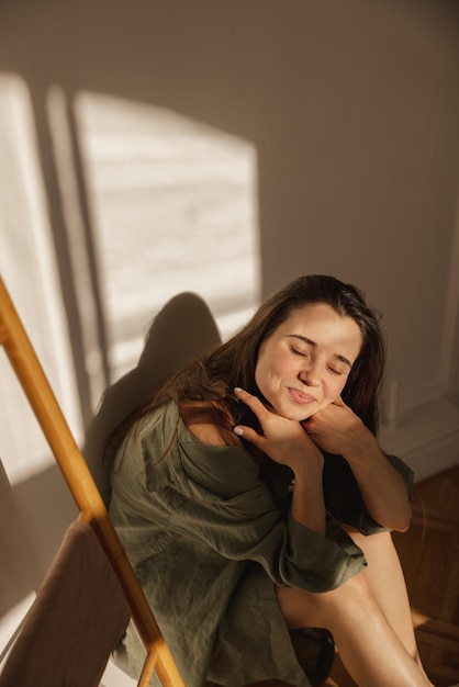 Free photo attractive caucasian young girl smiles modestly at camera closing her eyes from sun brunette sits in light room against white wall leisure lifestyle and beauty concept