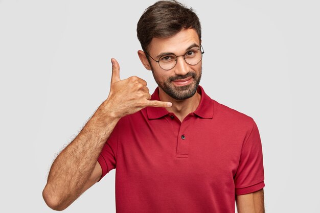 Attractive Caucasian young male in casual clothes imitates telephone conversation, keeps hand near ear as if holding mobile phone, has confident facial expression, isolated over white wall.