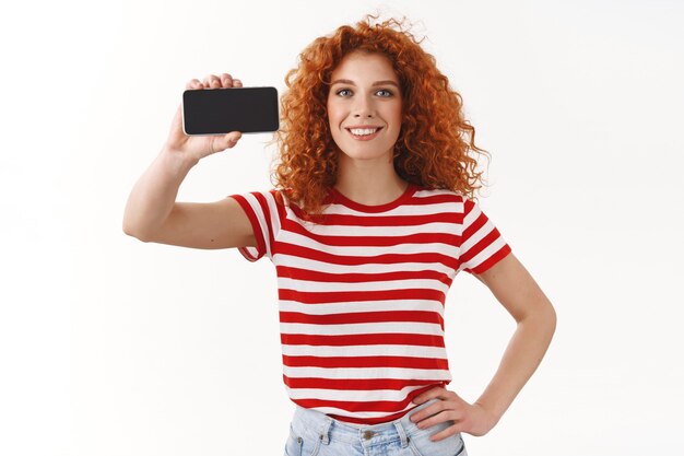 Attractive charismatic ginger girl curly hairstyle blue eyes hold hand waist showing smartphone display horizontal game promo smiling broadly advice cool app use