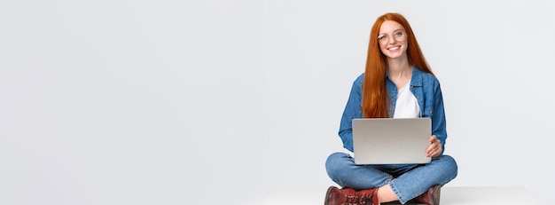 Foto gratuita la femmina caucasica rossa carismatica attraente si siede sul pavimento con le gambe attraversate che tengono i giri del computer portatile w