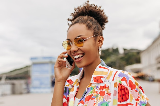 Attractive charming darkskinned lady in colorful stylish shirt and orange trendy sunglasses smiles and talks on phone outside