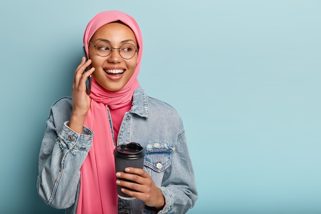 Attractive cheerful Arabian woman has telephone conversation with close friend, holds disposable cup of coffee, looks away