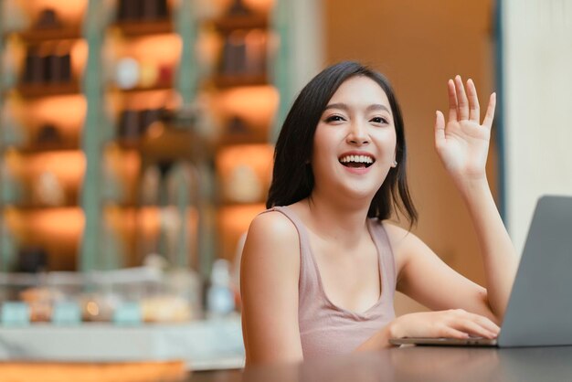 Attractive cheerful asian female adult gesture hand wave to say hi to her friend walking in cafe restaurantasian woman hand hold coffee cup relax casual in cafe