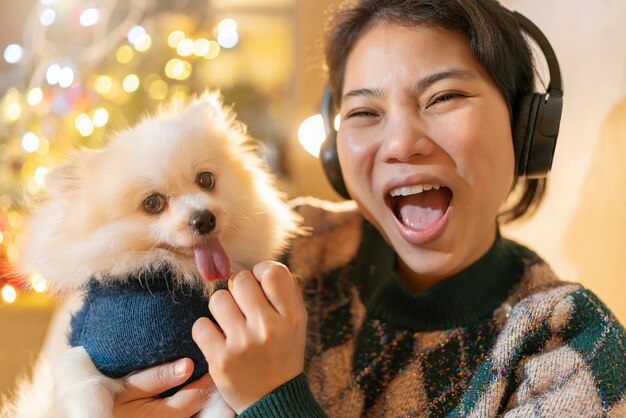 Attractive cheerful asian female wear winter cloth hand hold lapdog with sweater christmas costume enjoy talking photo selfie smartphone with hapiness smiling with christmas tree and lights bokeh