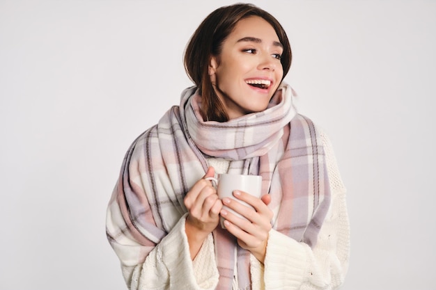 Бесплатное фото Привлекательная жизнерадостная девушка с уютным шарфом, держащая чашку с теплым напитком, счастливо отводящая взгляд на белом фоне