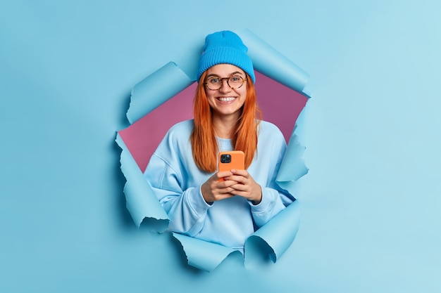 Привлекательная веселая женщина с рыжими волосами держит современные текстовые сообщения типа смартфона, любит серфить в социальных сетях, носит синюю шляпу и джемпер.