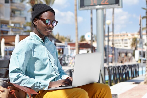 Attractive cheerful young African American male freelancer dressed in stylish wear sitting on urban bench with laptop computer on his lap and using free wireless internet connection for remote work