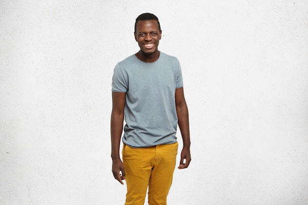 Free photo attractive cheerful young dark-skinned student in grey t-shirt and mustard jeans posing at white blank wall, smiling happily, enjoying nice time indoors after lectures at college