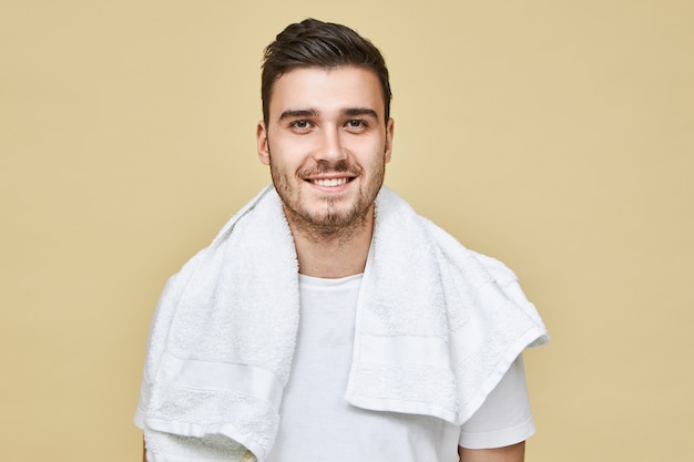 首の周りに無精ひげと白いタオルを持った魅力的な陽気な若いヨーロッパ人男性は、仕事の前の朝にバスルームで顔を剃るつもりで広く笑っています