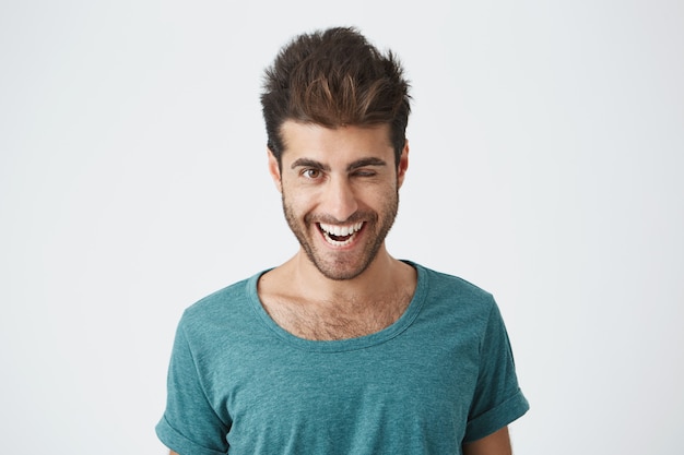 Attractive and cheerful young man with beard and stylish haircut wearing blue t-shirt blinking his eyes with pleasure having happy expression. Facial expressions and people emotions concept